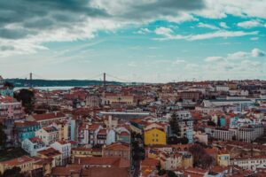 Red-roofed houses across Lisbon with the 25 de Abril Bridge crossing the Tagus river under a cloudy blue sky