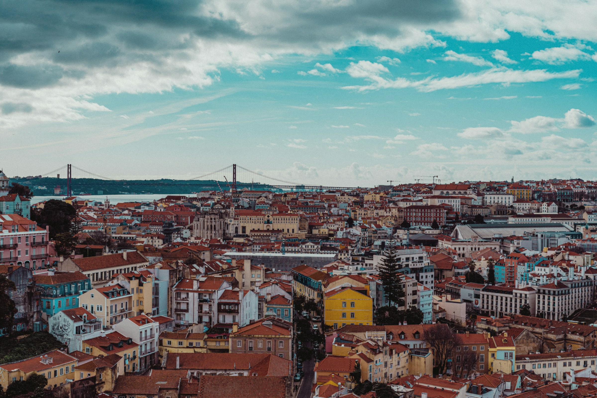 Red-roofed houses across Lisbon with the 25 de Abril Bridge crossing the Tagus river under a cloudy blue sky